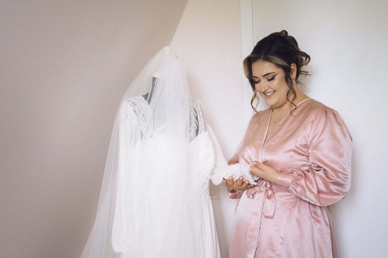 Bride in pink satin robe admiring her white wedding dress and veil hanging on the wall before the ceremony.