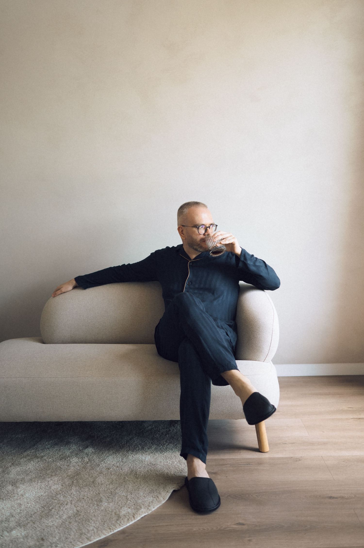 Bald man with glasses in dark outfit sipping drink while relaxing on modern beige sofa in minimalist room.