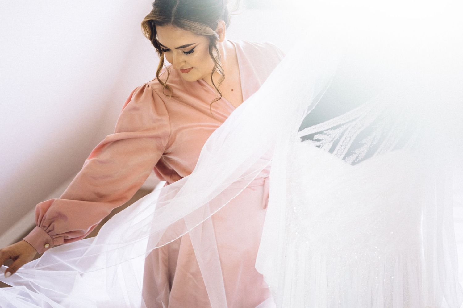 Bride in pink dress holding white veil in bright light.