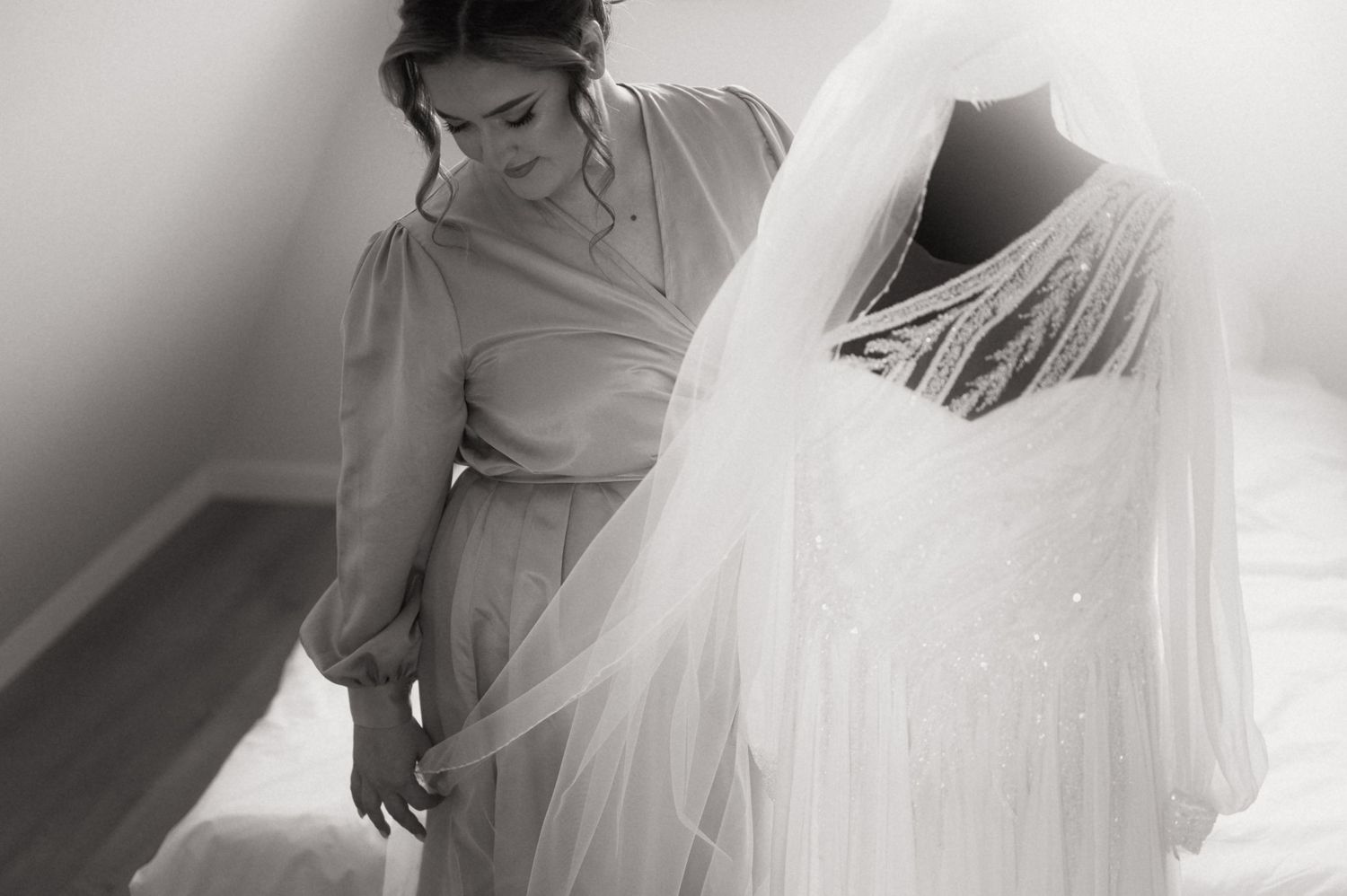Black and white photo of a bride admiring her elegant wedding dress on a mannequin with a flowing veil.