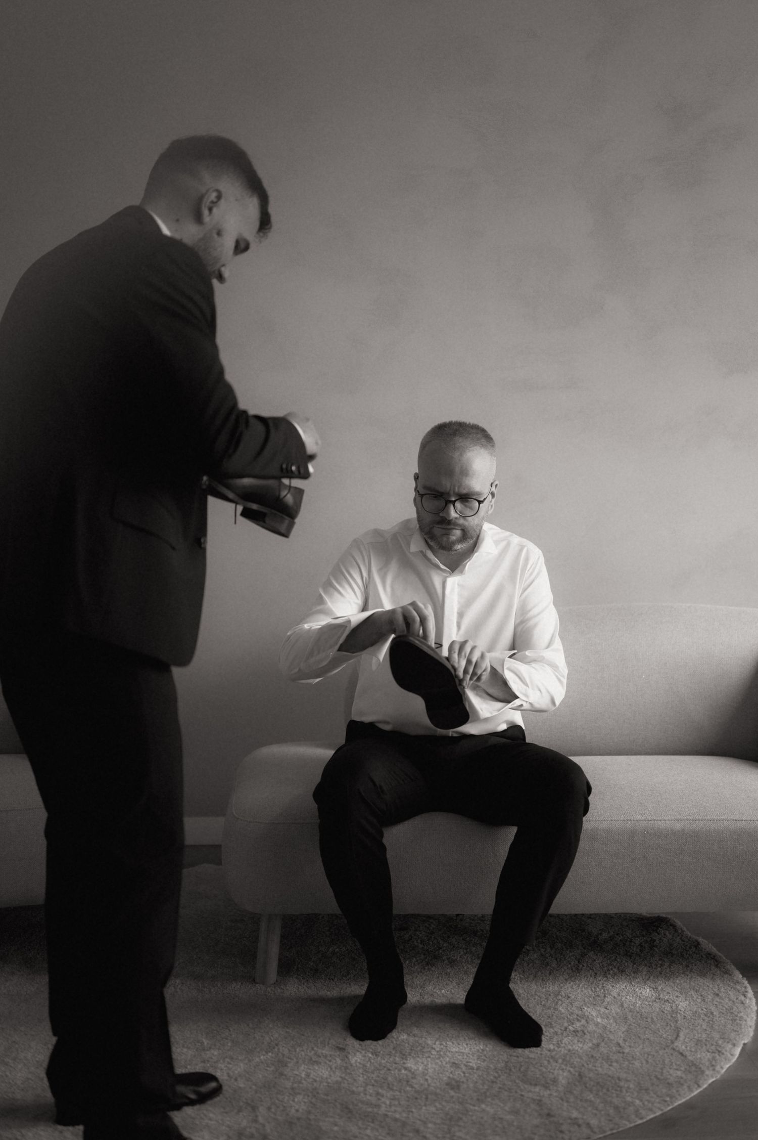 Black and white photo of two men in formal attire, one seated putting on shoes while another stands holding items.