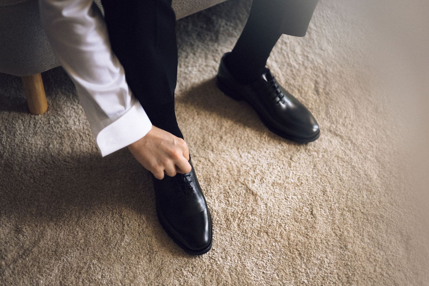 Man in formal attire putting on black dress shoes while getting ready for a wedding or formal event.