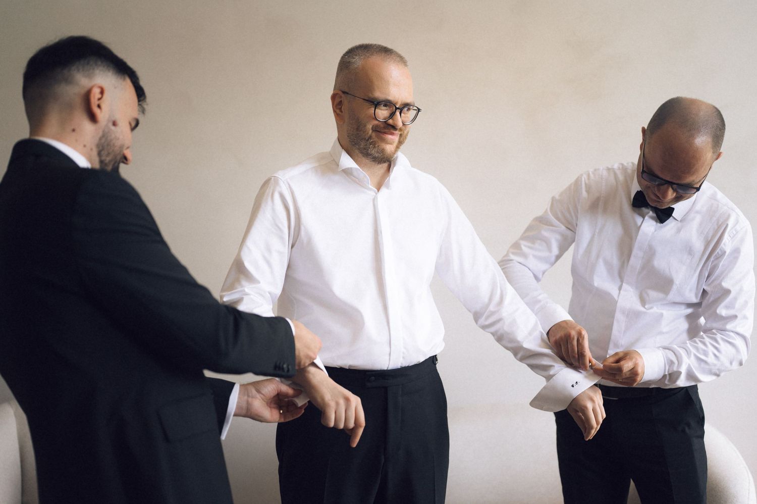 Three men in formal black and white attire adjusting their cufflinks and bow ties before a wedding ceremony.