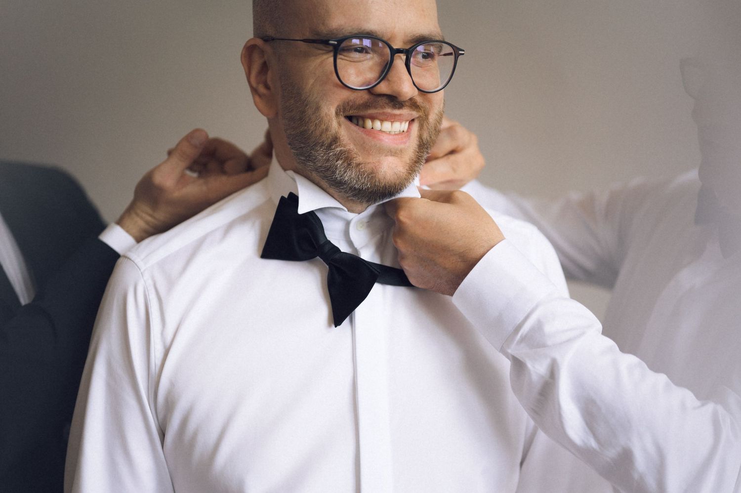 Smiling bald man in white shirt adjusting black bow tie with glasses, assisted by someone behind him.