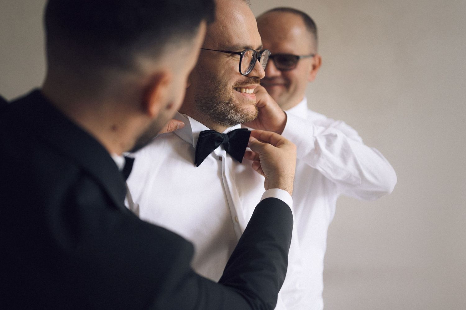 Two groomsmen helping a groom adjust his black bow tie while dressing for a wedding ceremony.
