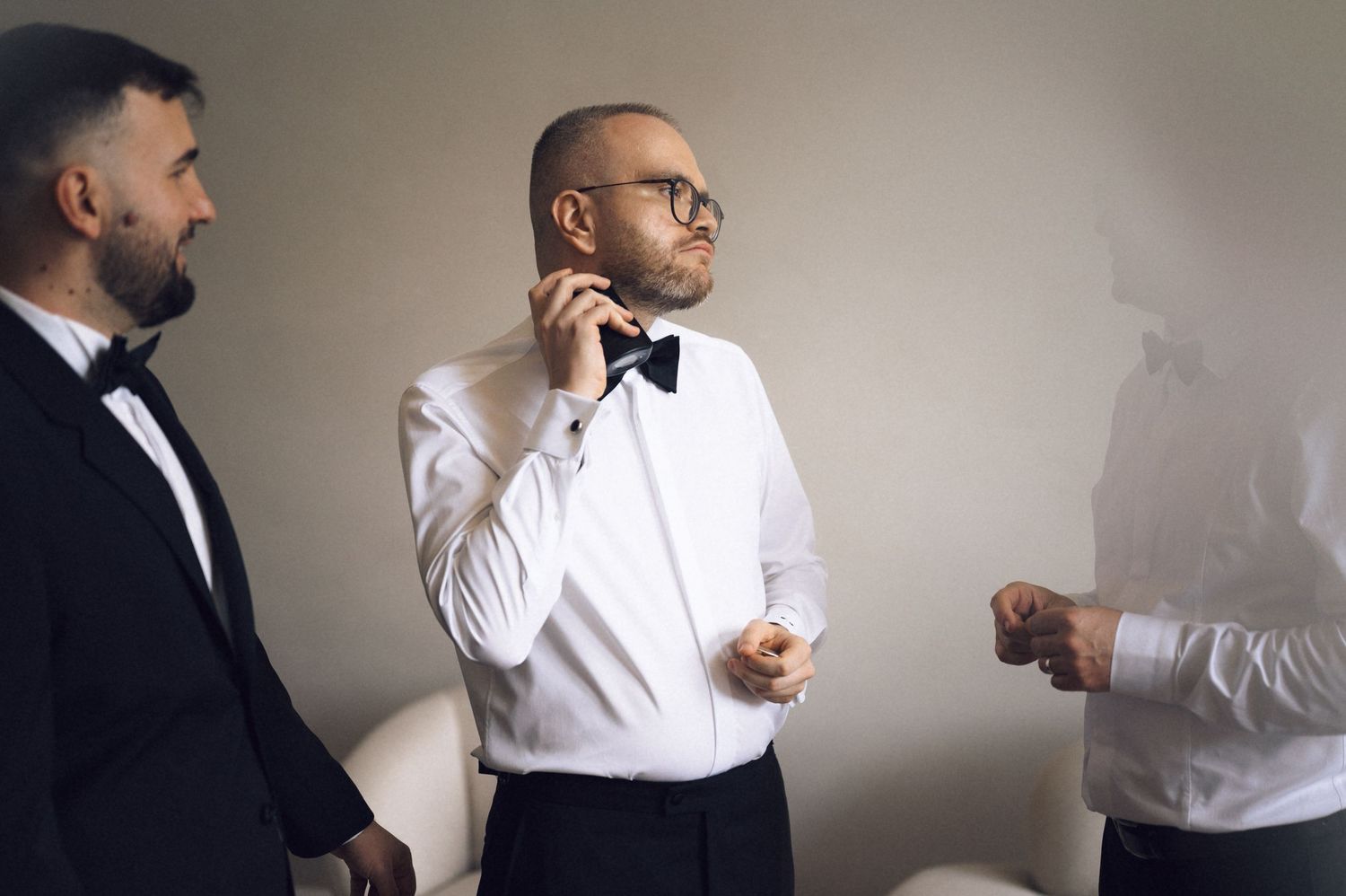 Three men in formal tuxedos with bow ties prepare for a wedding ceremony indoors.