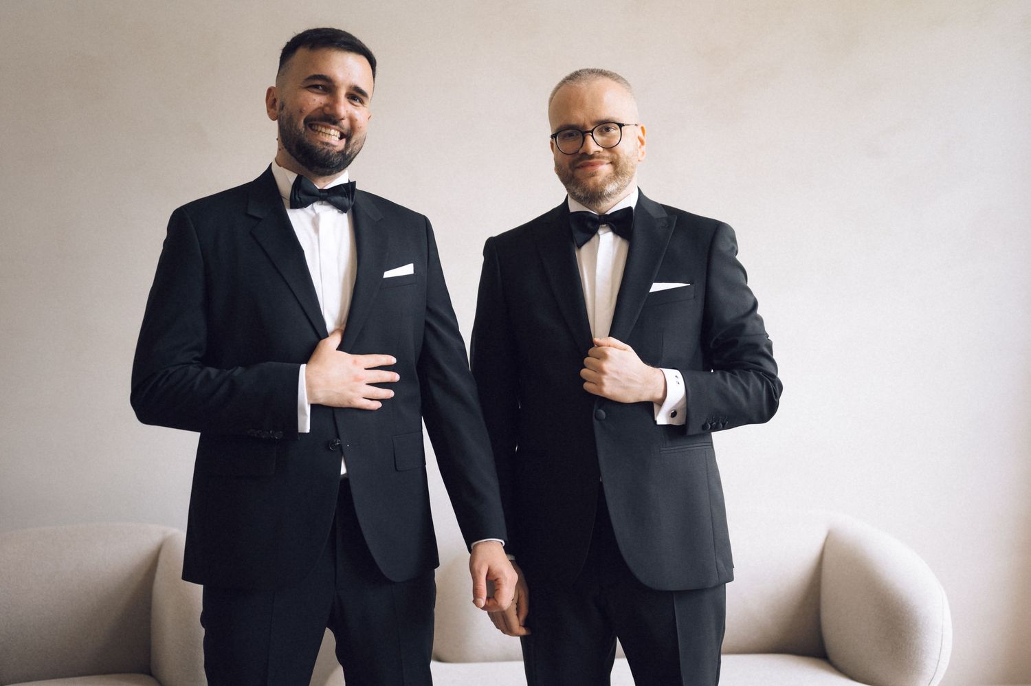 Two men in matching black tuxedos with bow ties smile while adjusting their jackets in an elegant white room.