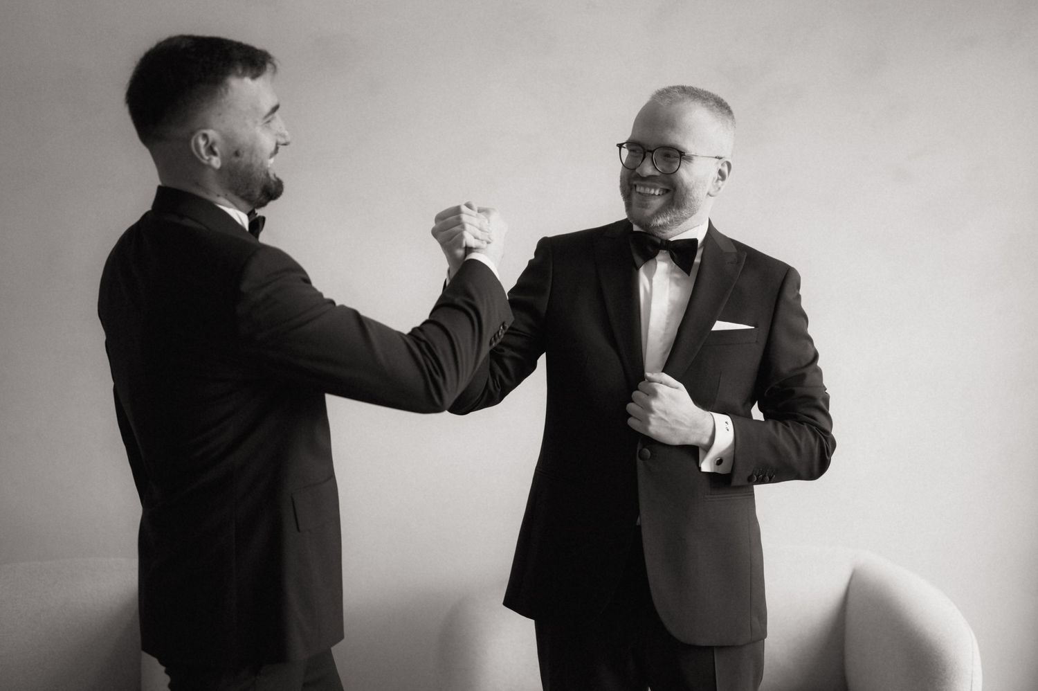 Two men in tuxedos share a fist bump while getting ready for a formal event in a black and white photo.
