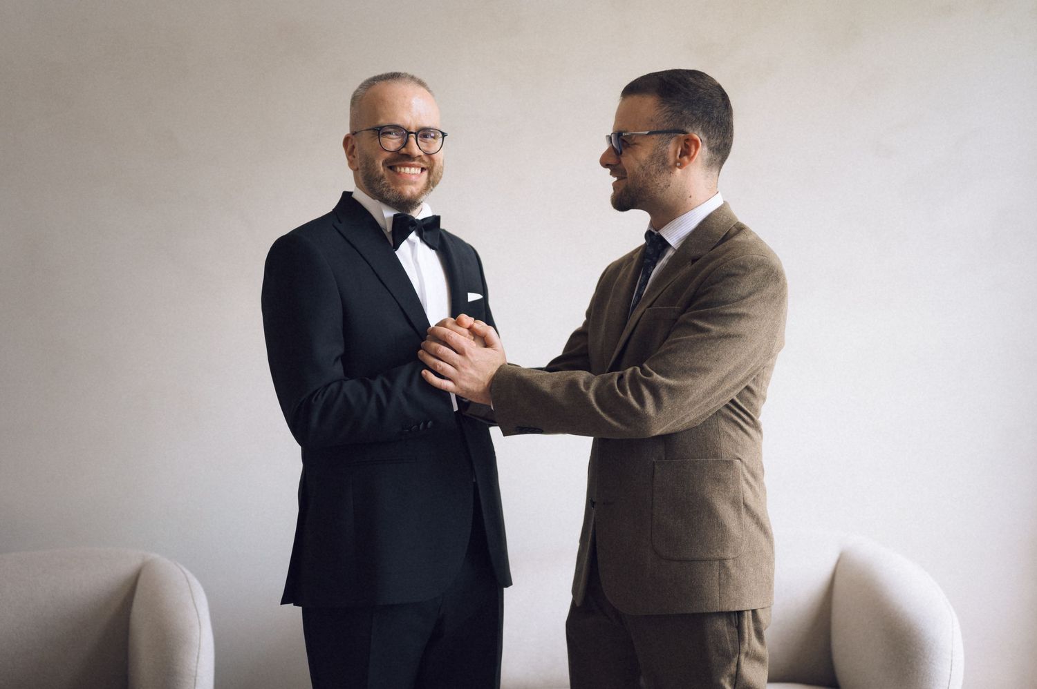 Two men in formal suits smiling and shaking hands at a wedding celebration indoors.