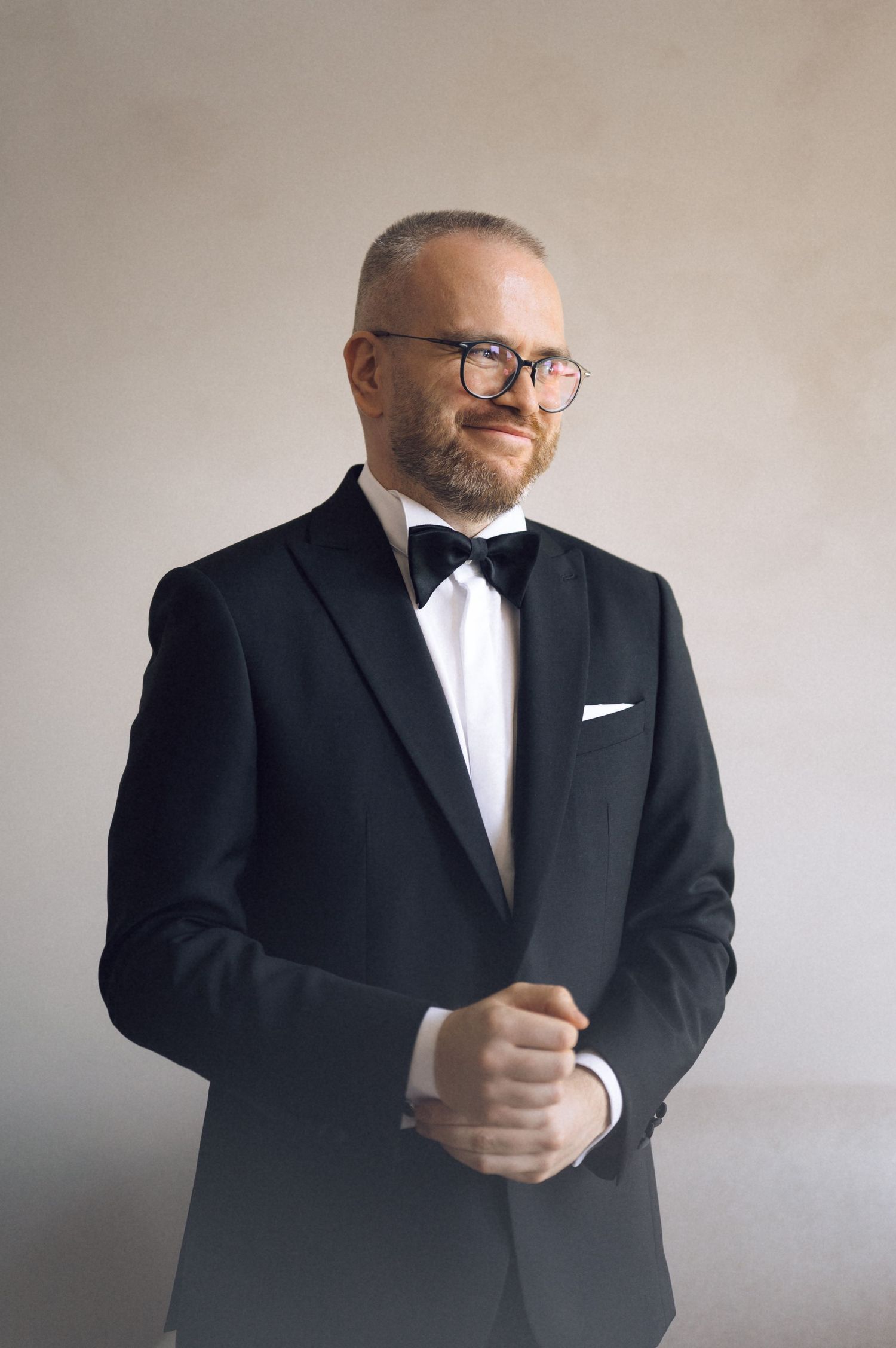 Man in black tuxedo with bow tie and glasses posing confidently in a formal portrait setting.
