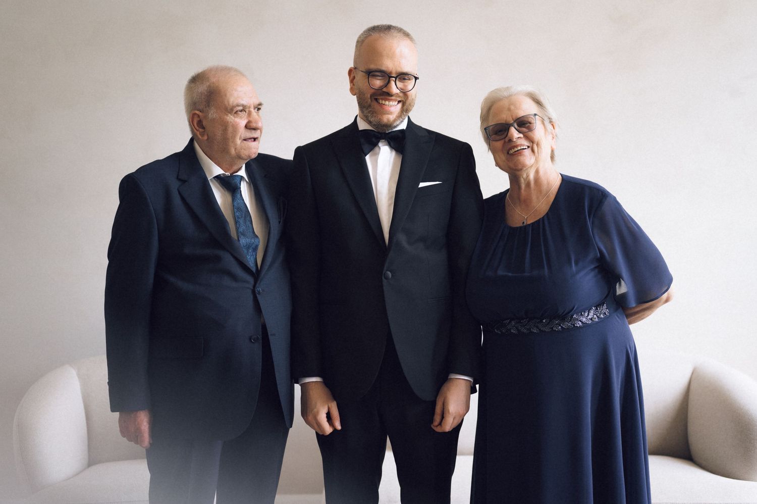 A groom in a tuxedo poses with an older couple in navy formal wear, smiling together at an elegant wedding venue.