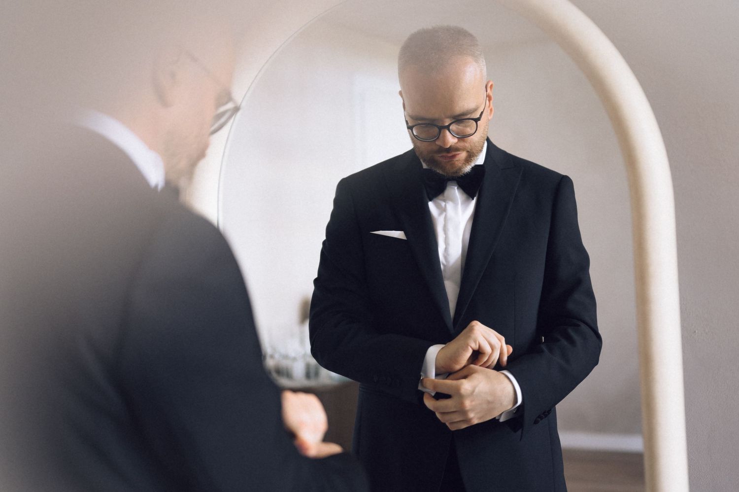 Bald man in black tuxedo and bow tie adjusting cufflinks before mirror, elegant wedding preparation photo.