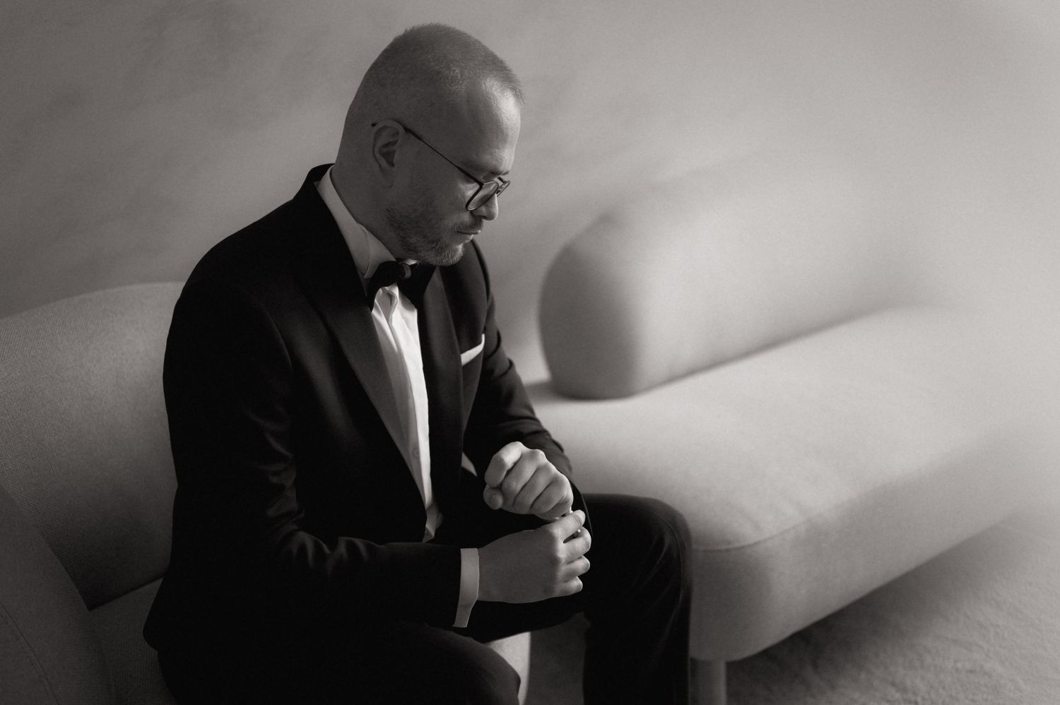 Man in black tuxedo and bow tie sits pensively on white sofa, black and white portrait photography.