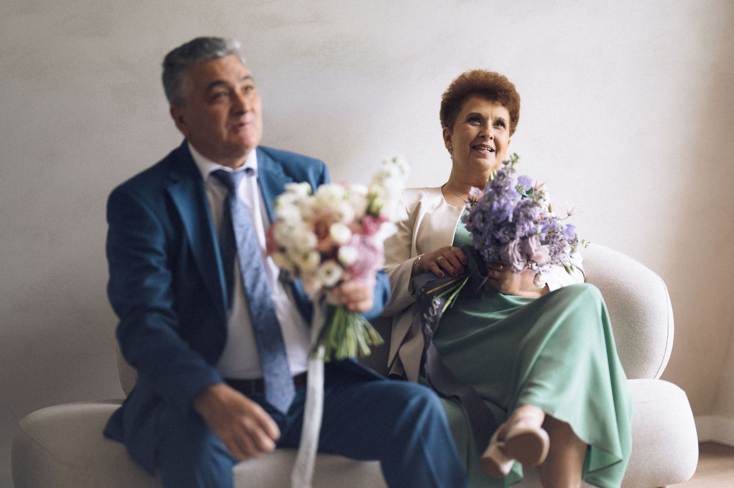 Elderly couple sitting on white sofa at wedding, man in blue suit holding bouquet, woman in green dress with purple flowers.