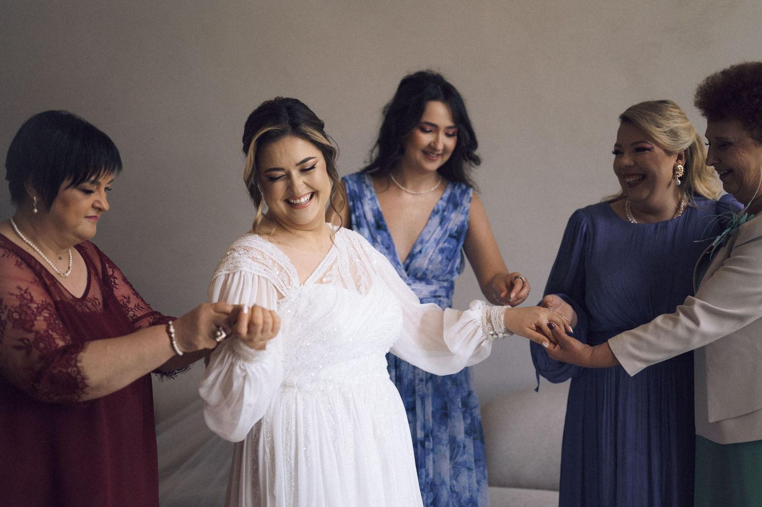 Bride in white gown smiles as bridesmaids in blue and red dresses help her prepare for her wedding day.