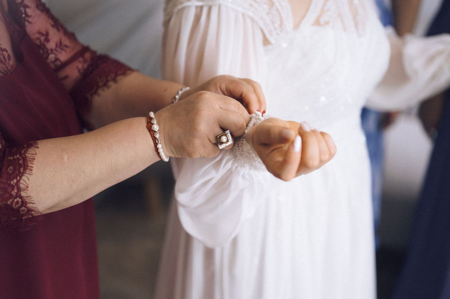Close-up of hands fastening a button on a white wedding dress, with a woman in a red lace dress assisting the bride.