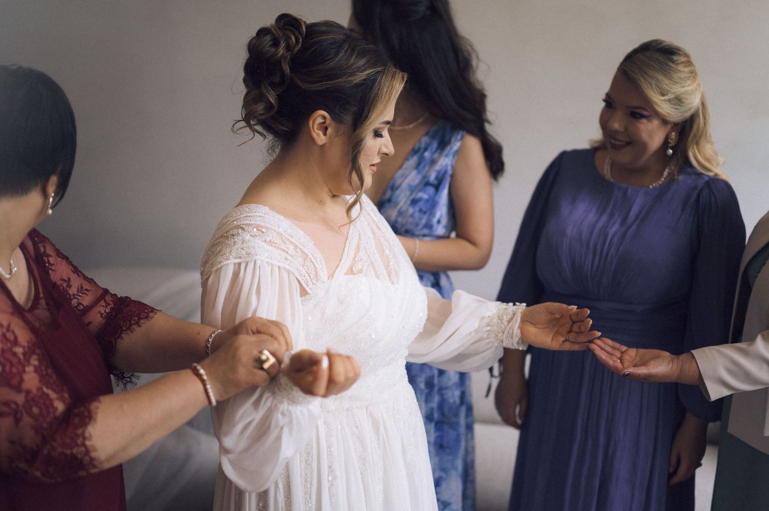 Bride in white dress being helped by bridesmaids in blue and red during wedding preparation.