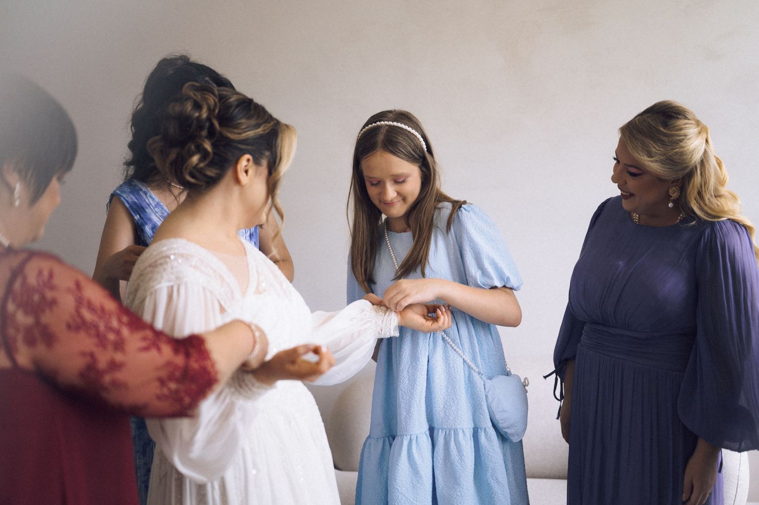 Women in elegant dresses gather closely, examining something delicate during a bridal preparation moment.