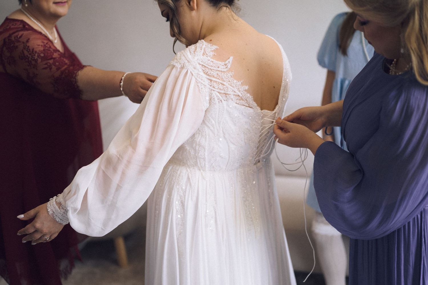 Bridesmaids helping bride button up her white lace wedding dress with flowing sleeves before the ceremony.