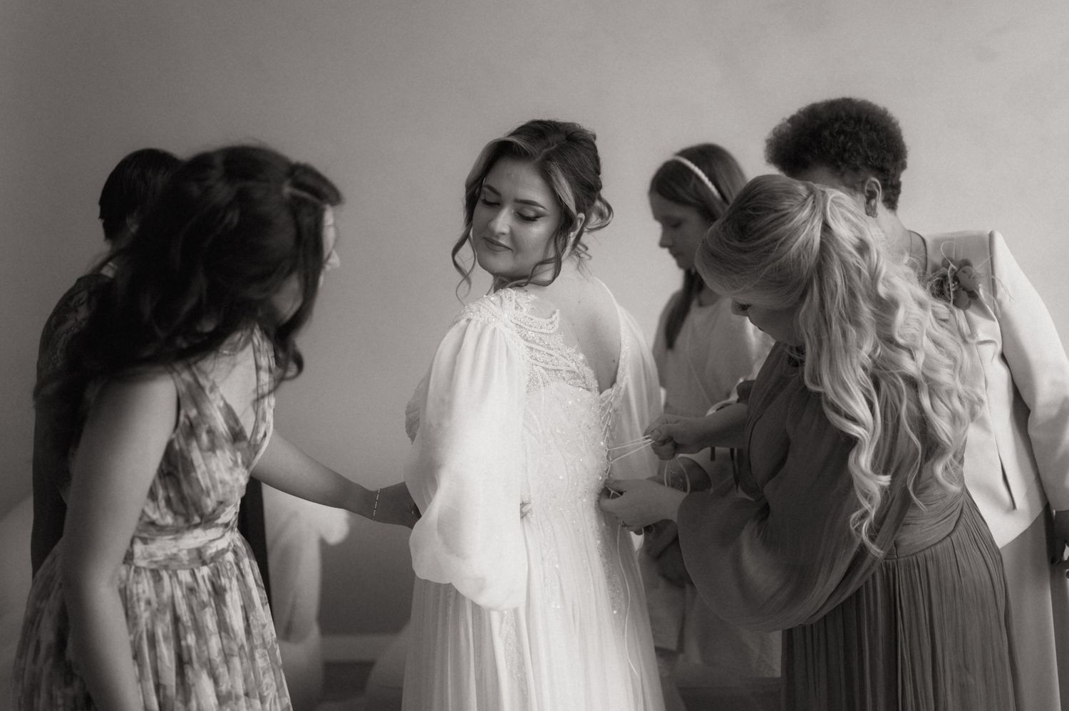 Black and white photo of a bride in a white gown being helped by bridesmaids while getting ready for her wedding.