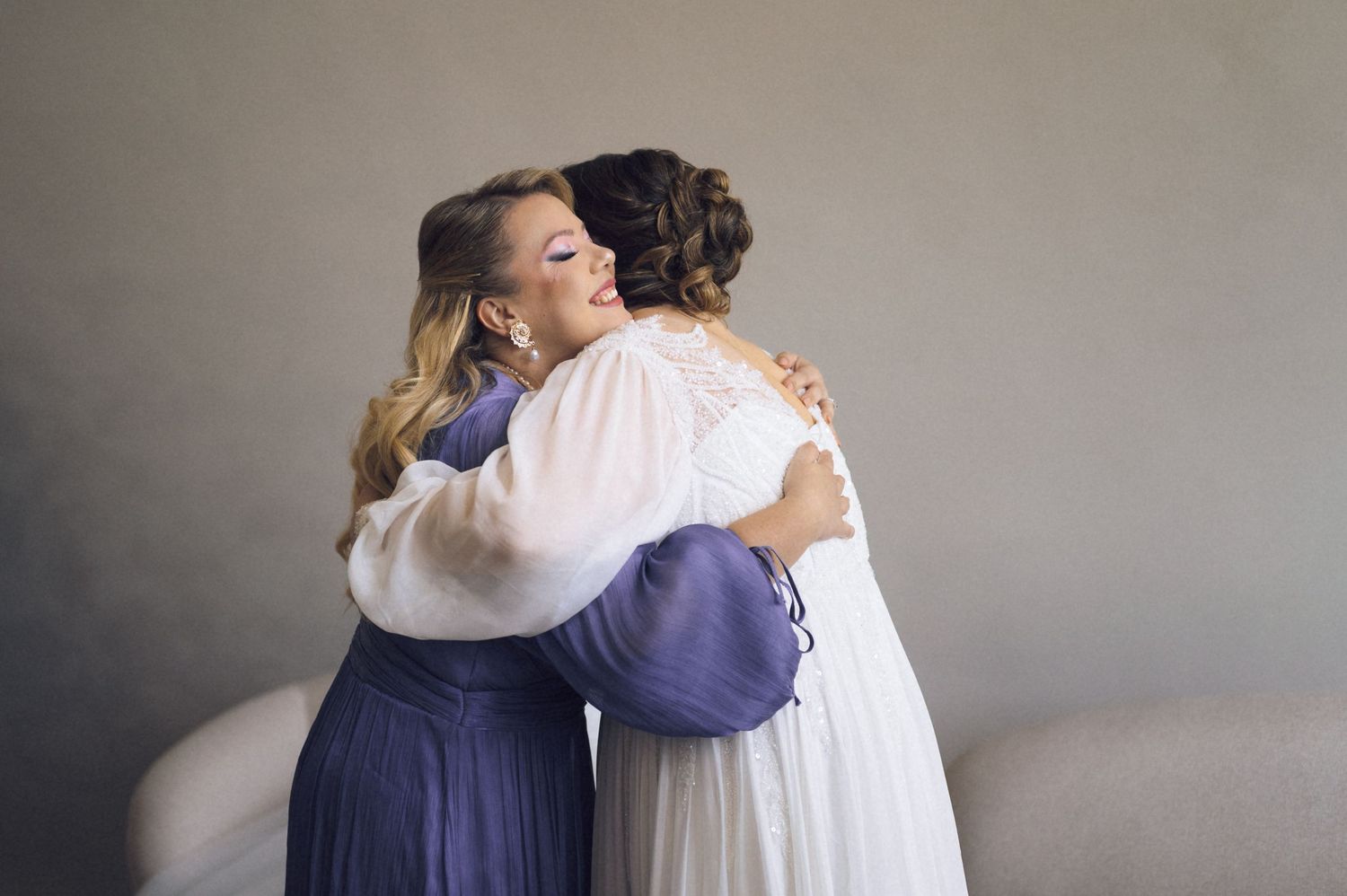 Two women in elegant attire share a warm embrace, one in a white bridal gown and the other in a purple dress.