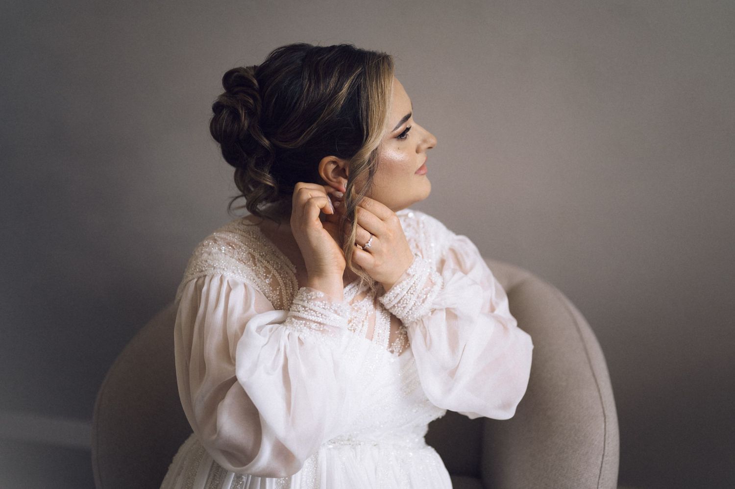 Bride in white lace dress adjusting earring while seated in elegant chair, soft natural lighting.