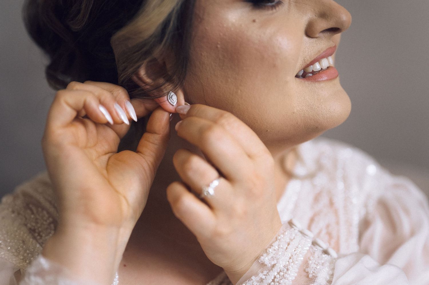Bride putting on elegant pearl and crystal earrings, wearing lace dress and diamond engagement ring on her wedding day.