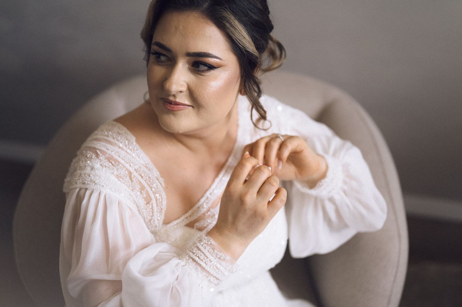 Bride in white lace gown adjusting her ring while seated in a gray chair, featuring elegant updo and blue eye makeup.