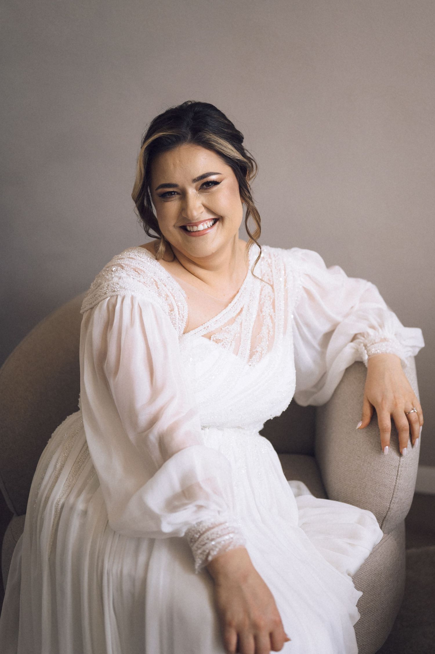 Smiling bride in a white long-sleeve lace wedding dress seated in a gray chair against a neutral background.