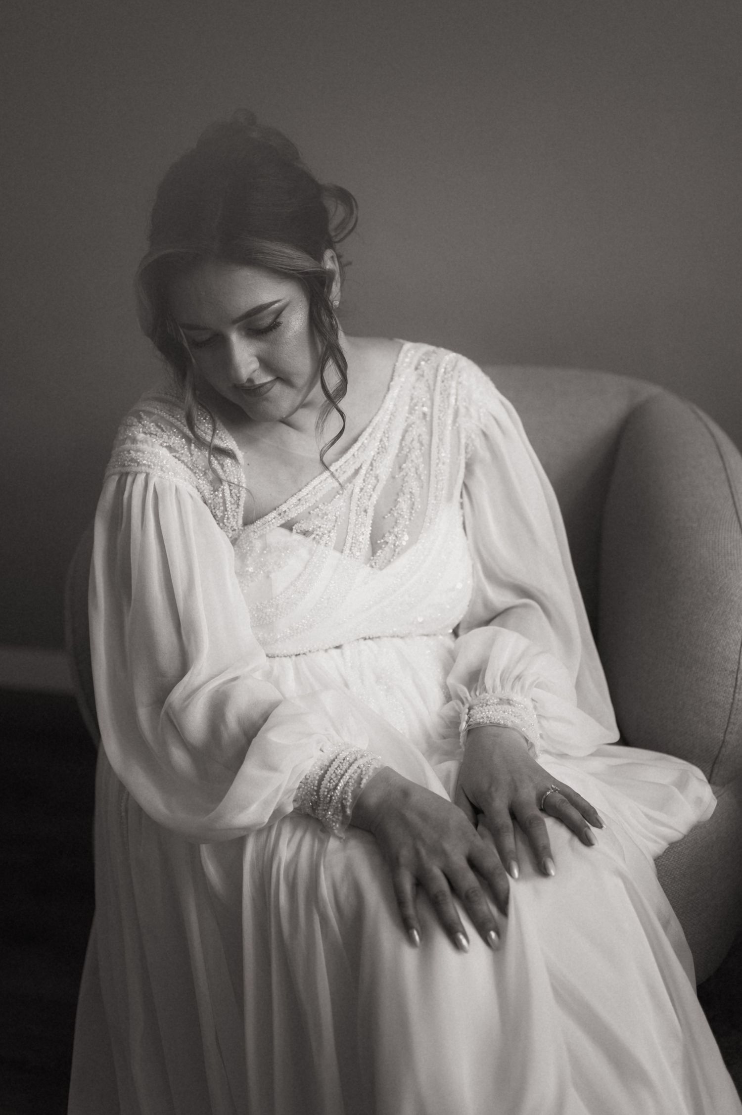Black and white photo of a bride in a flowing white lace gown sitting gracefully in a chair, looking down at her hands.