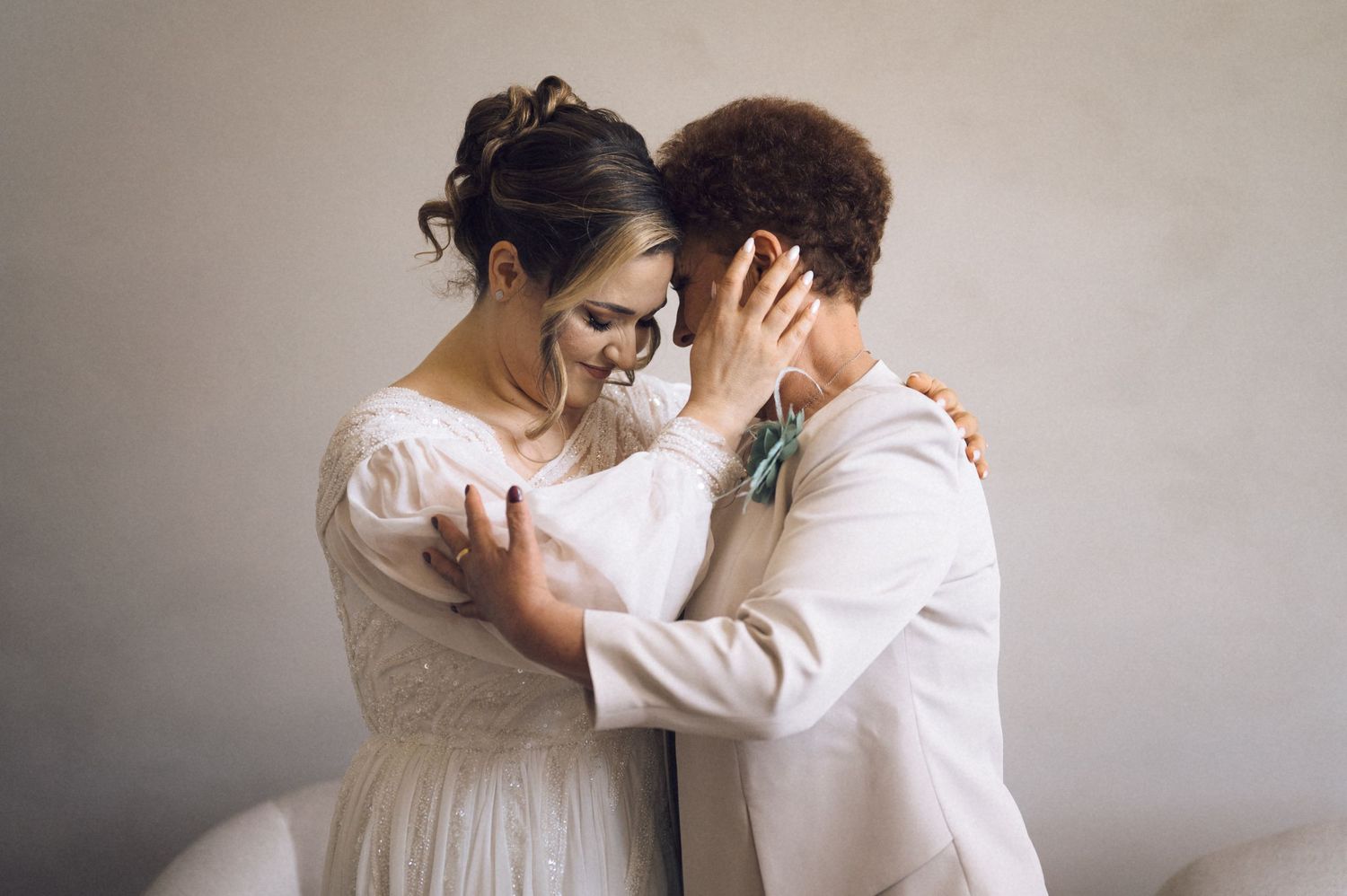 Two brides share an intimate moment in white wedding attire, one in a flowing gown, the other in a chic suit.