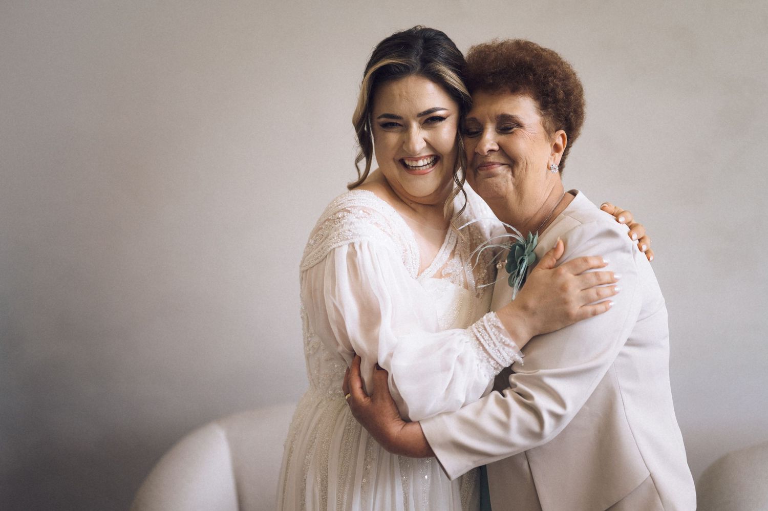 A bride in a white dress smiles while embracing an older woman in a beige suit at a wedding celebration.