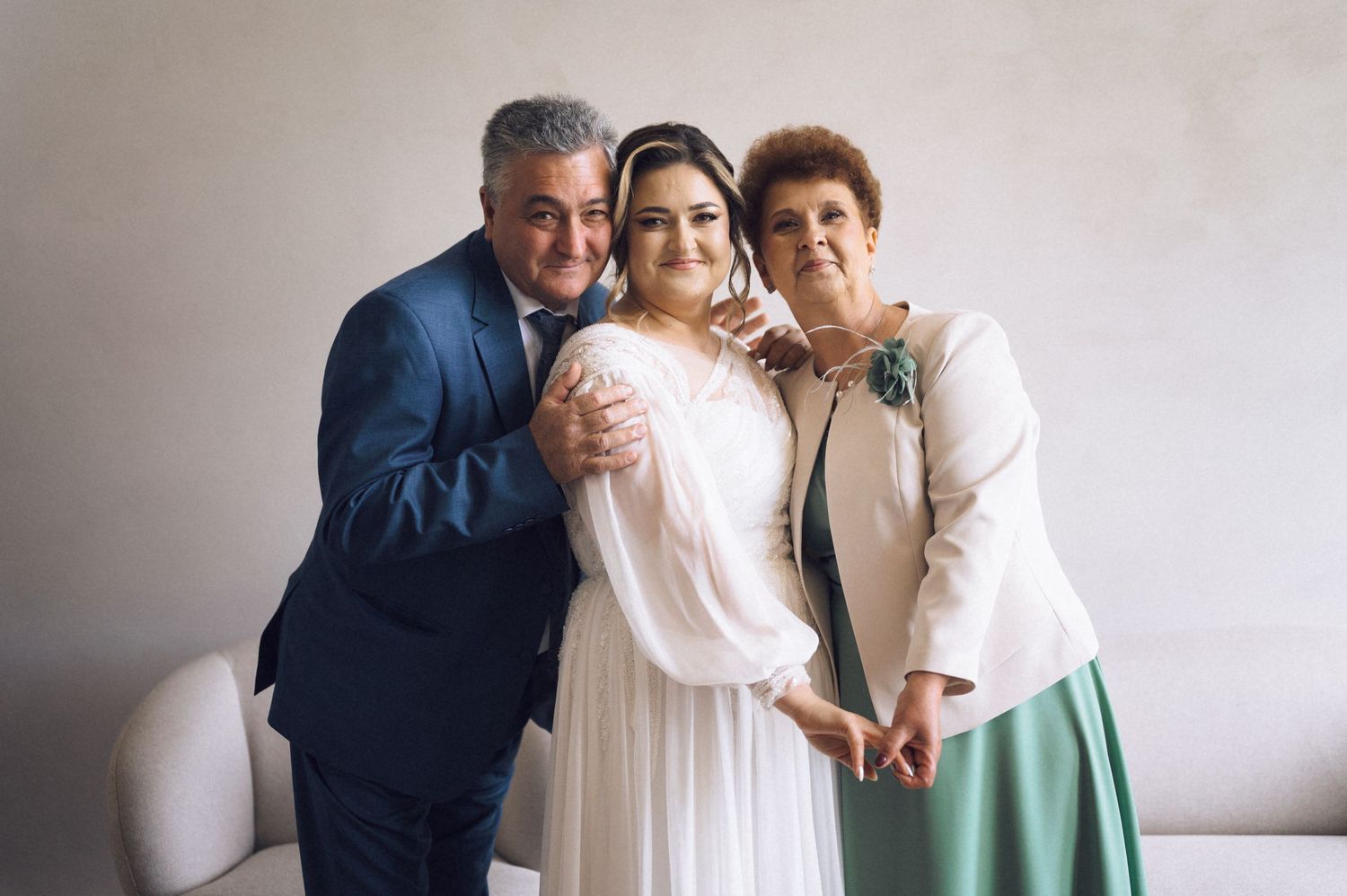 A bride in a white gown poses with her parents, a man in a blue suit and a woman in a white jacket.