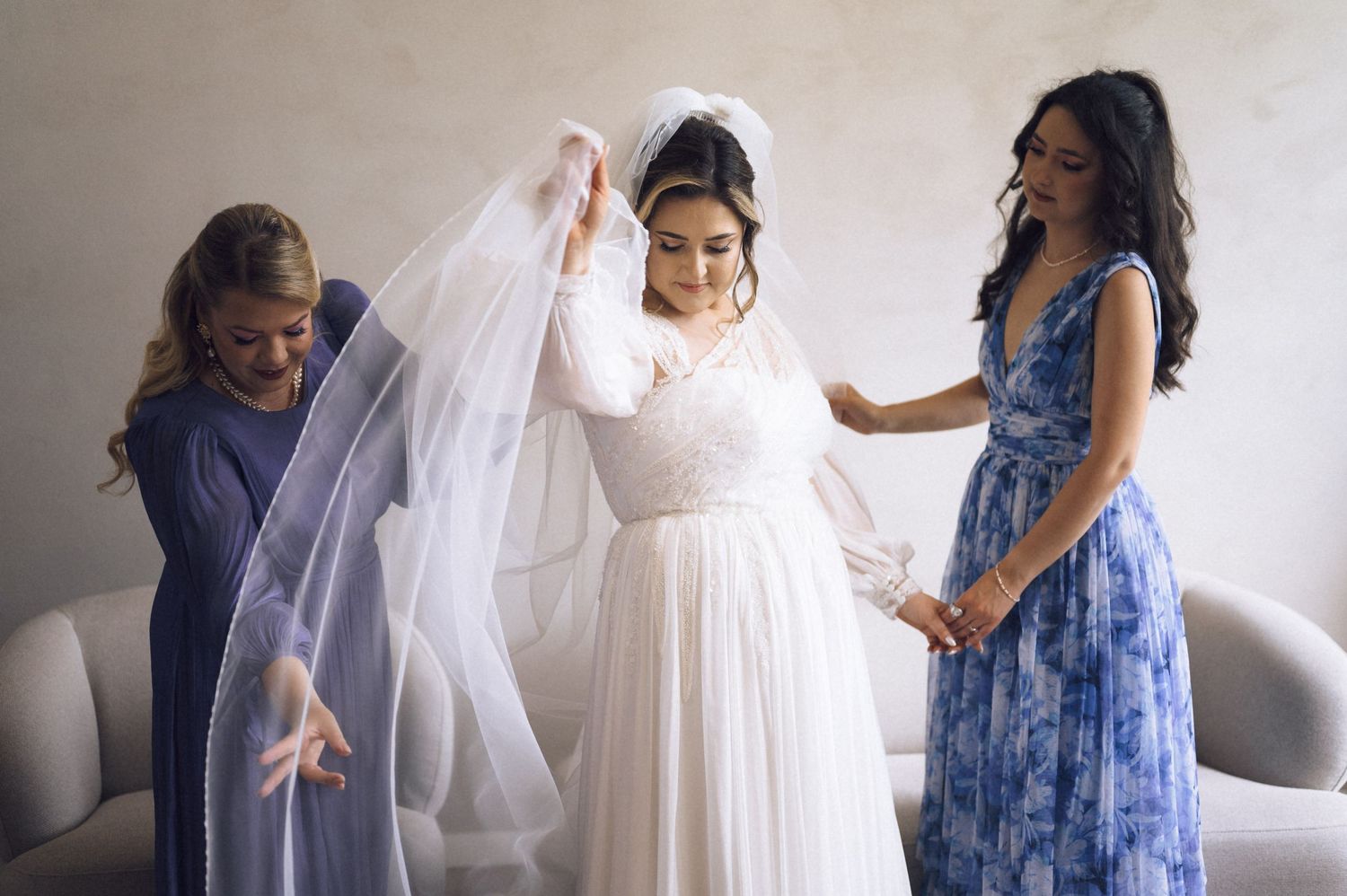 Bride in white lace gown and veil being helped by two bridesmaids in blue dresses during wedding preparations.