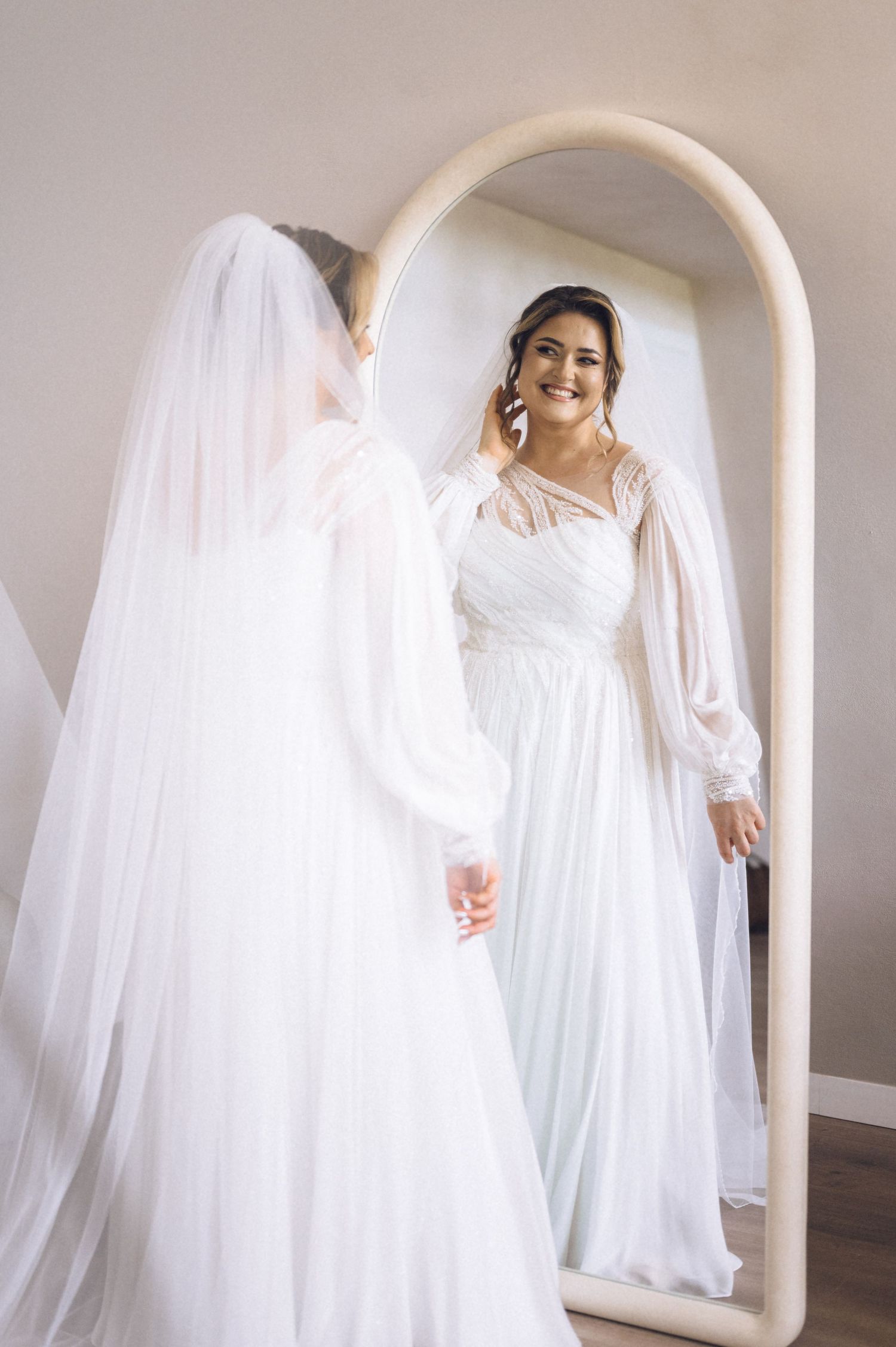 Bride in a white long-sleeve gown and veil smiles at her reflection in a large arched floor mirror.