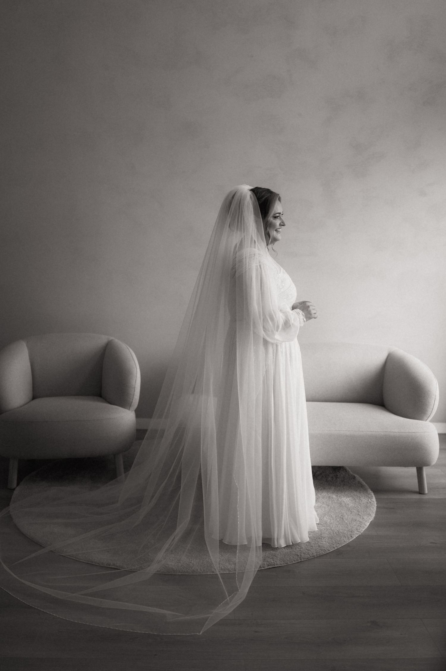 Black and white photo of a bride in a flowing white gown and long veil standing elegantly by a window.