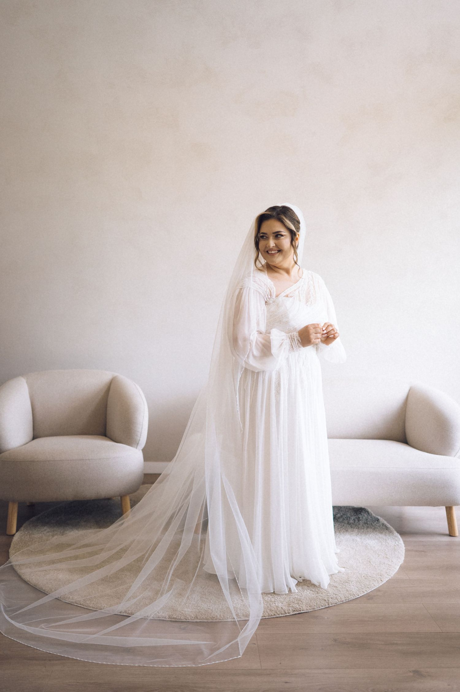 Bride in a white long-sleeve wedding dress with a flowing cathedral veil, standing in a minimalist room.