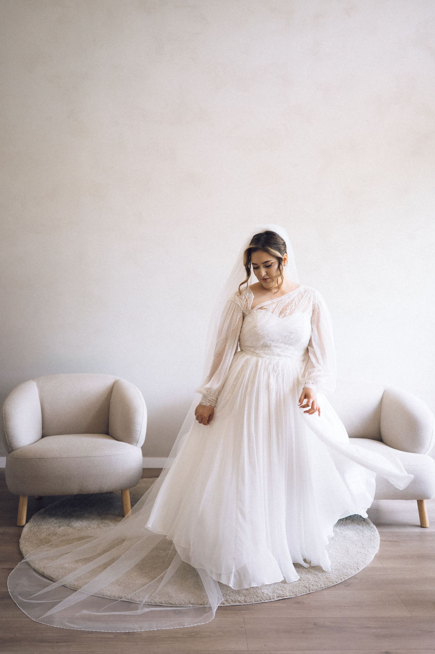 Bride in elegant long-sleeve white tulle wedding gown with veil, standing gracefully in a softly lit bridal suite.