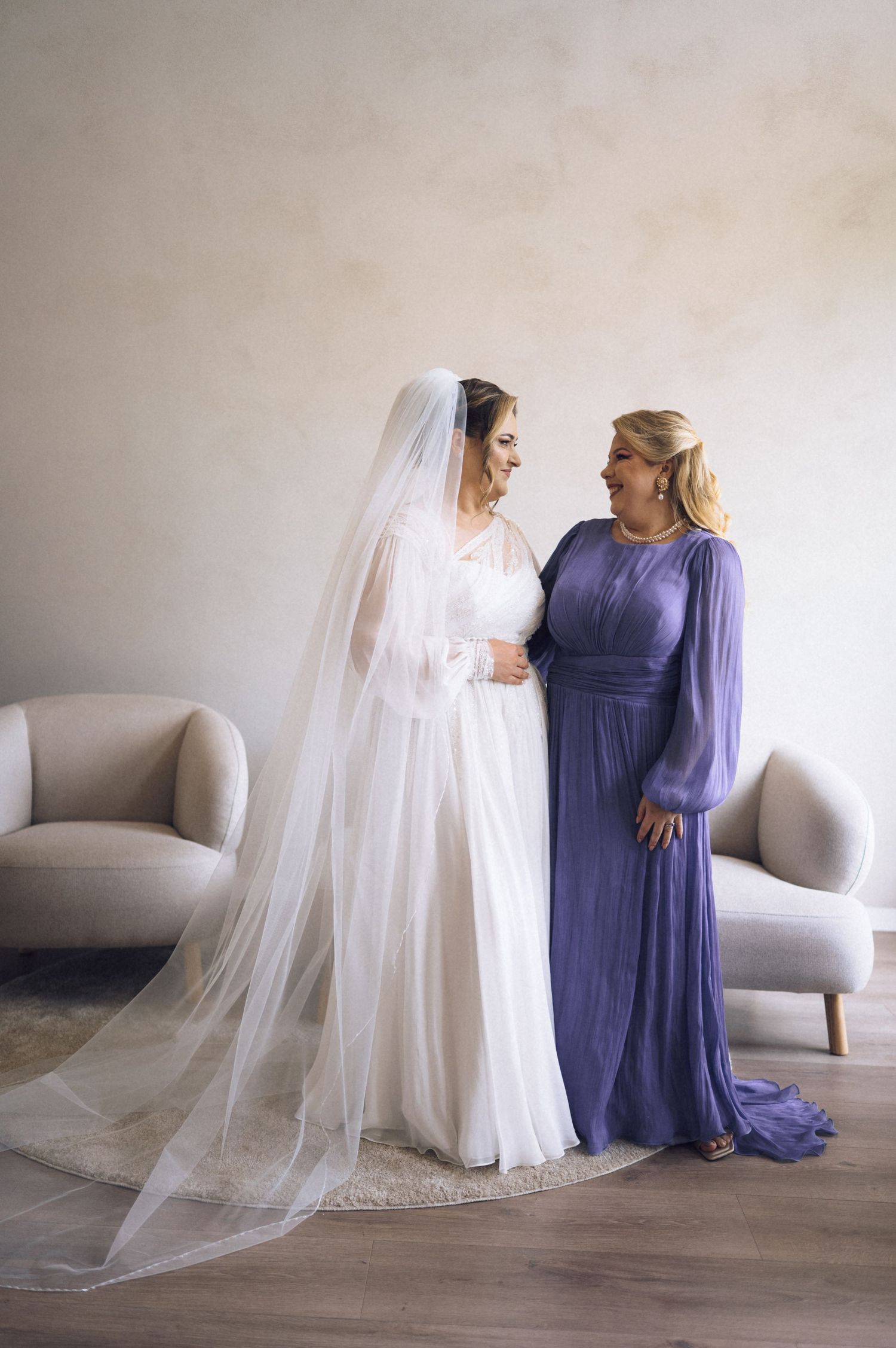 Bride in white gown and veil stands with woman in purple dress, sharing a joyful moment in an elegant room.