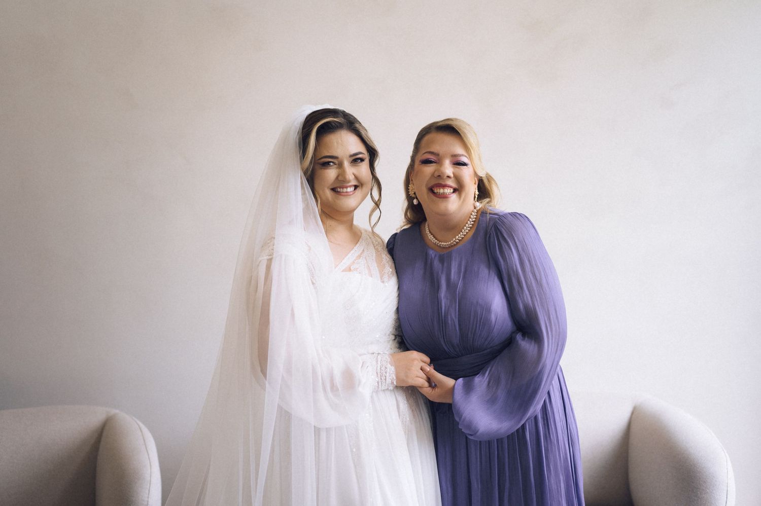 Bride in white gown and veil poses with woman in purple dress, smiling together at a wedding celebration.