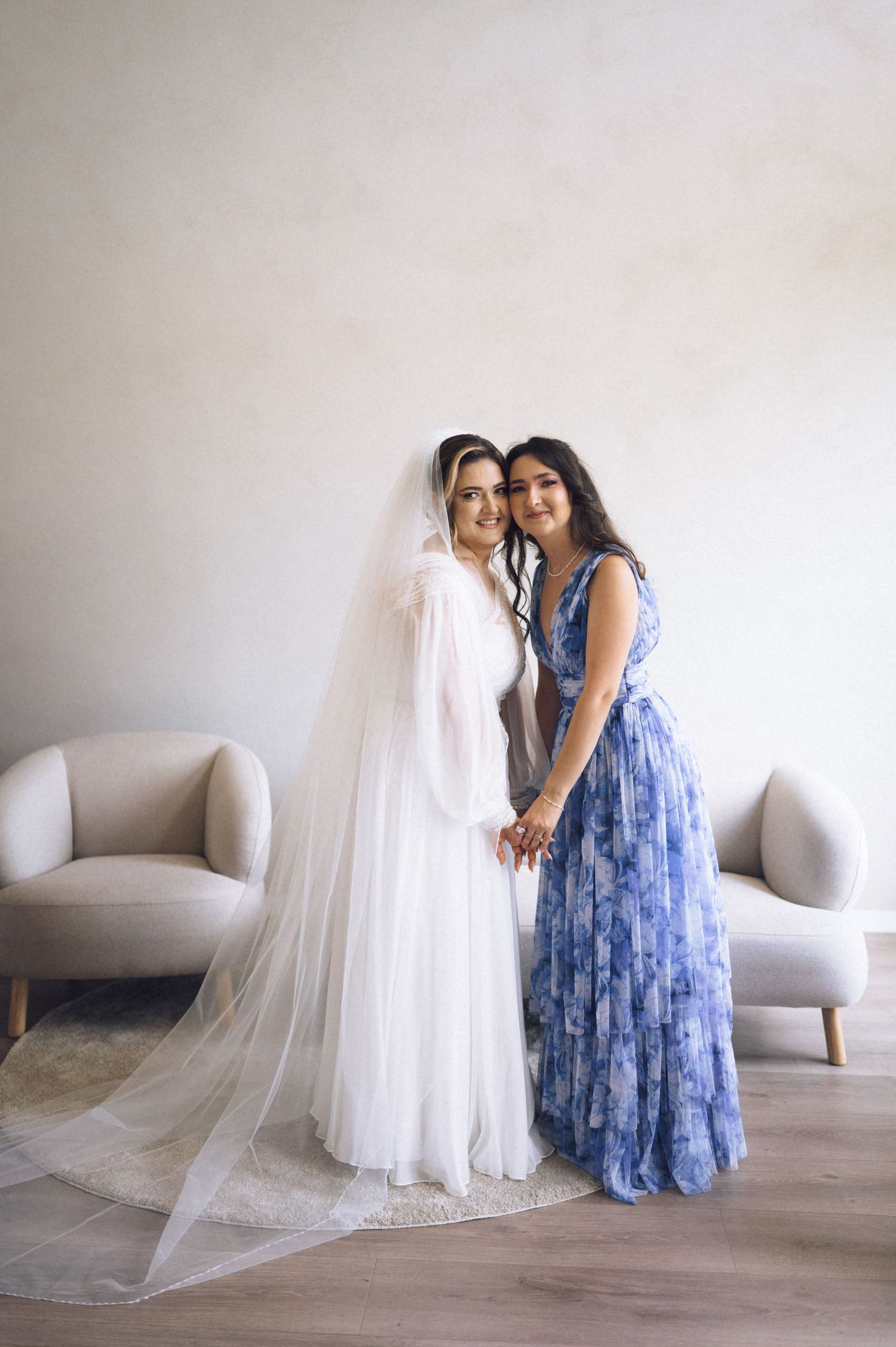 Bride in white gown with veil stands beside bridesmaid in blue floral dress, holding hands indoors.