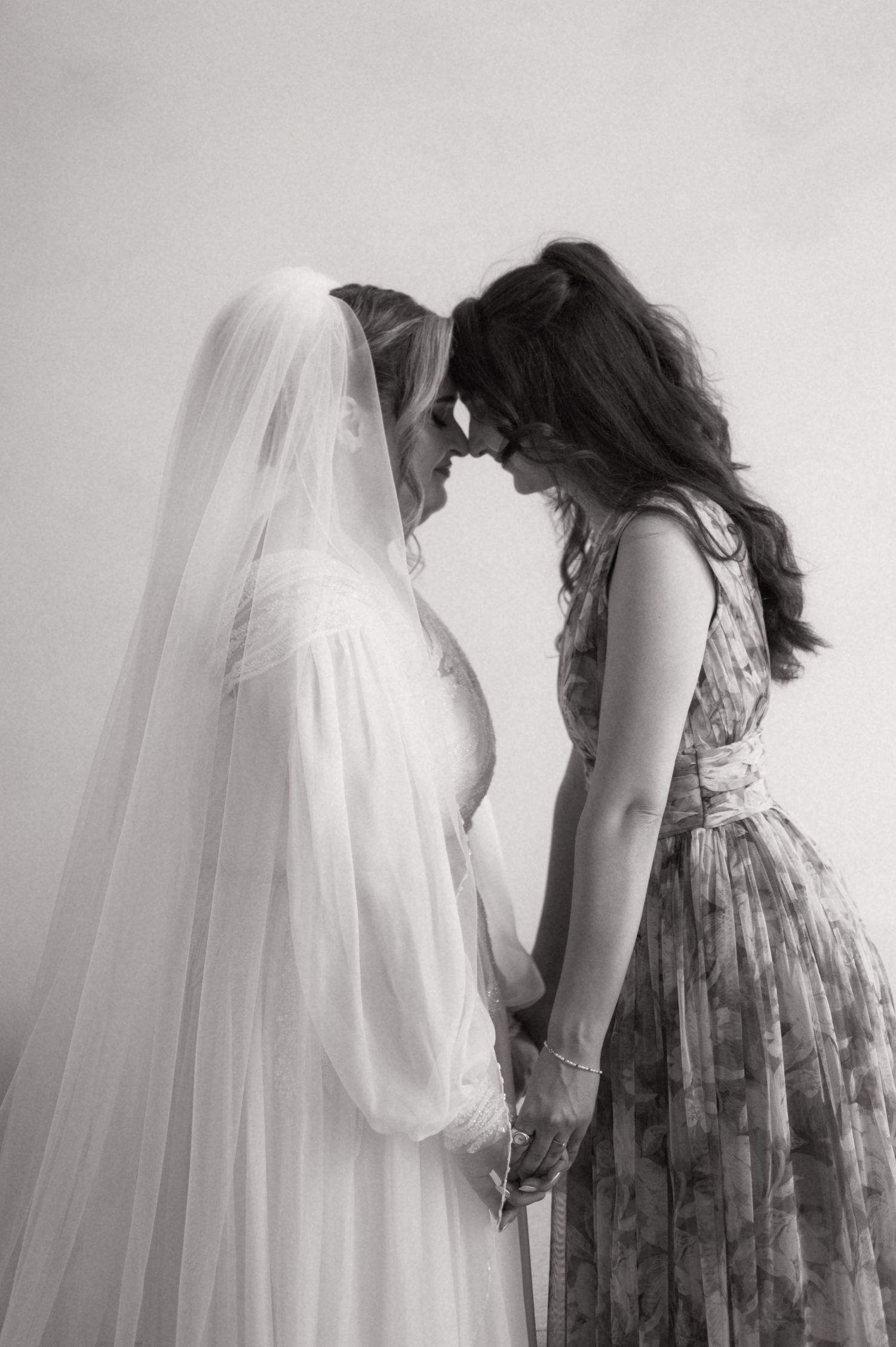 Black and white photo of a bride in veil holding hands and touching foreheads with a woman in floral dress.
