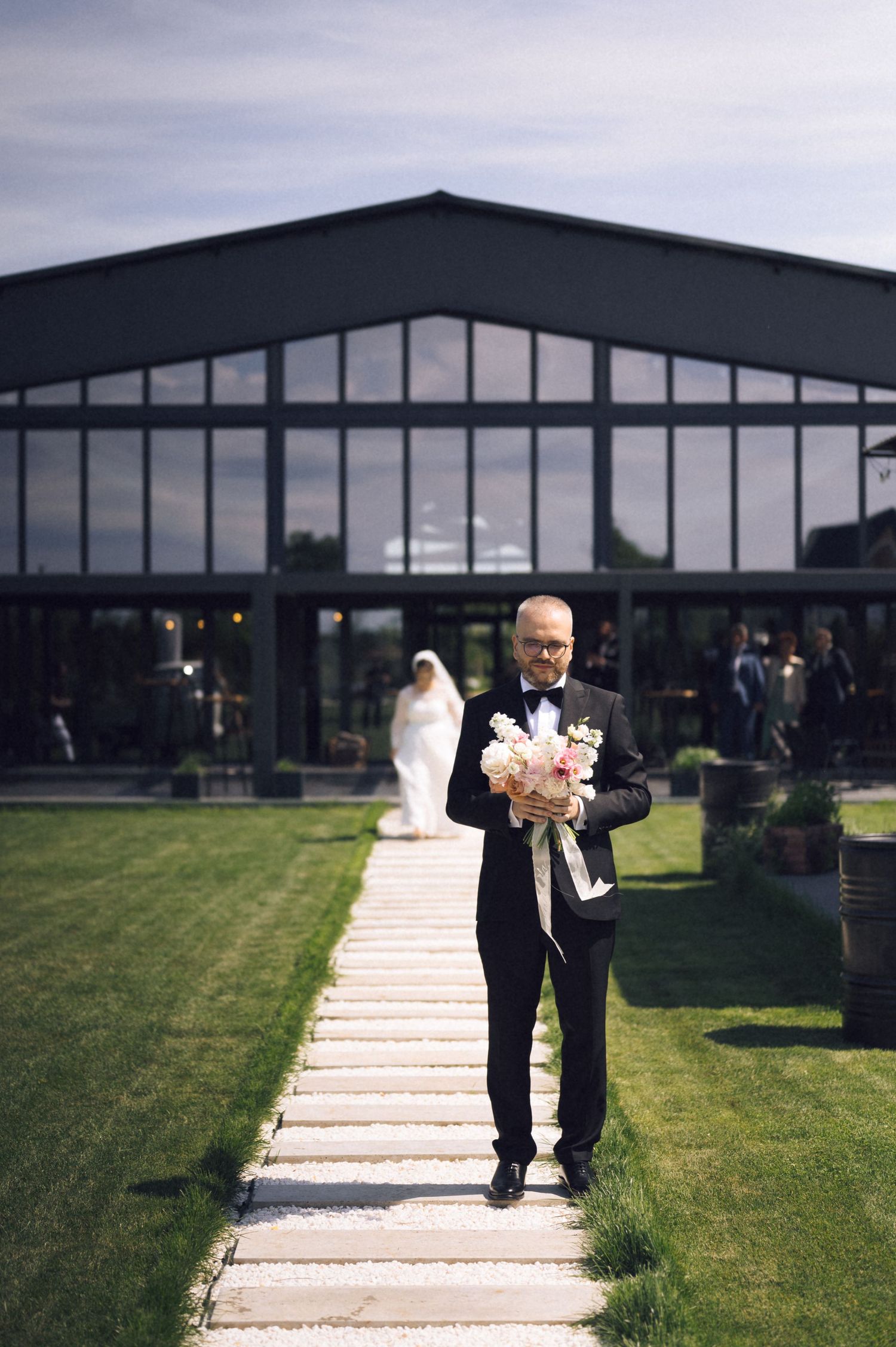 Man in tuxedo holding bridal bouquet walks stone path toward camera with bride in white gown visible in background.
