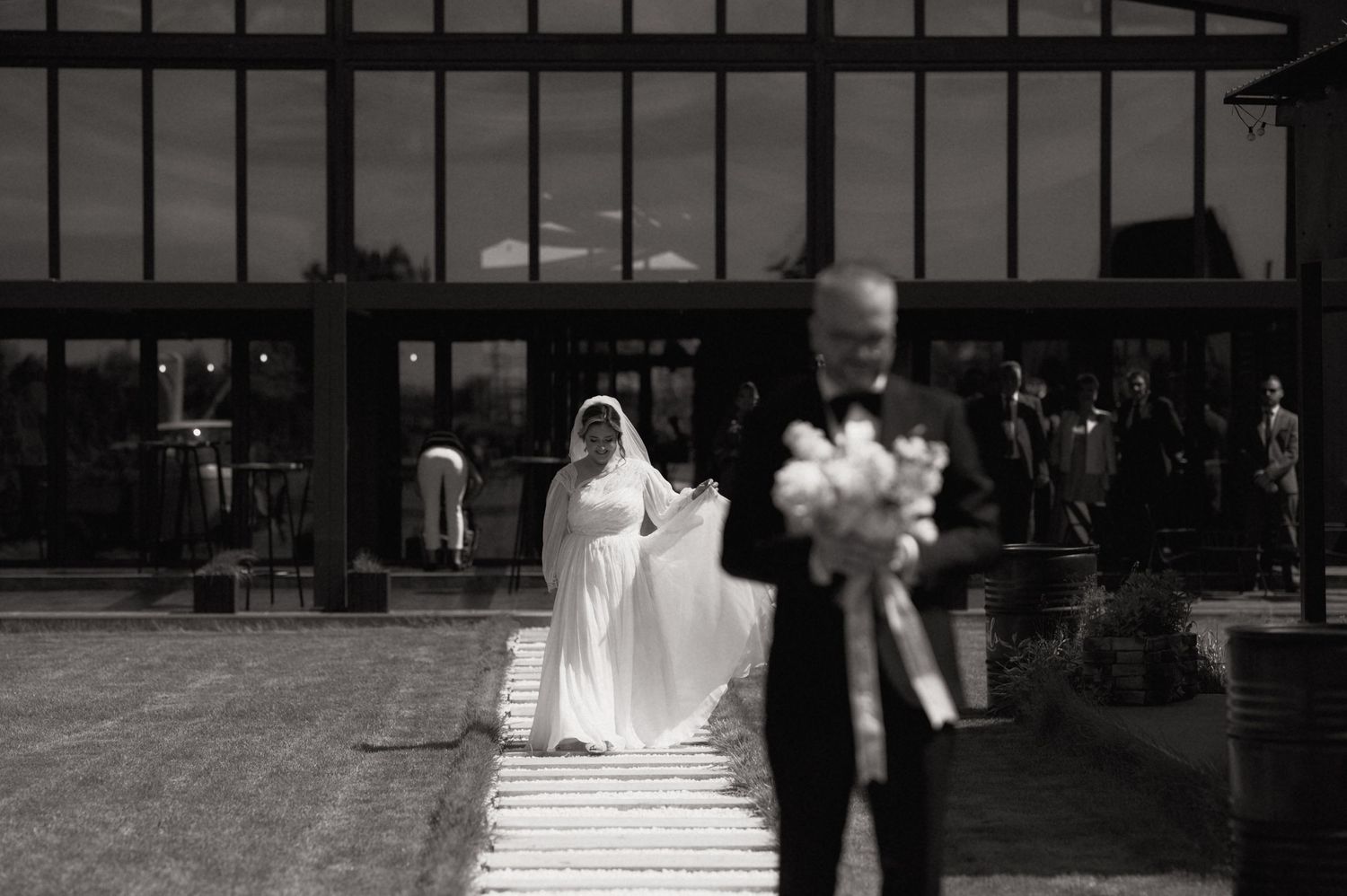 Black and white wedding photo of bride in white gown walking toward groom holding bouquet at elegant venue.