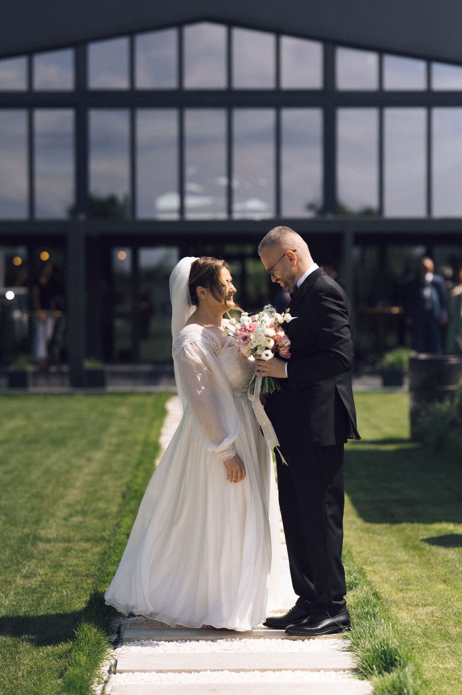 Bride in white gown and veil holds bouquet with groom in black suit, standing on garden path before large glass building.