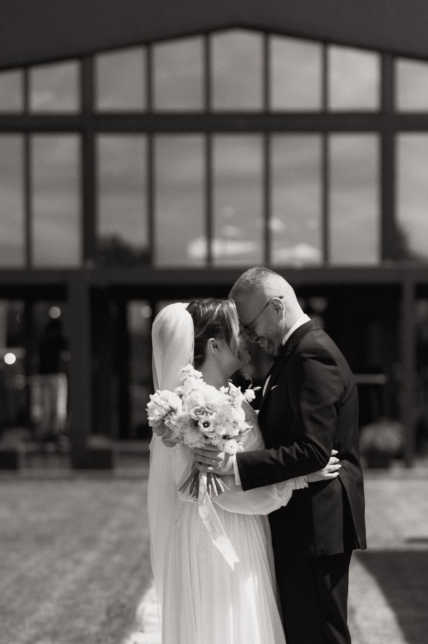 Black and white photo of bride and groom touching foreheads, sharing intimate moment outside modern venue.