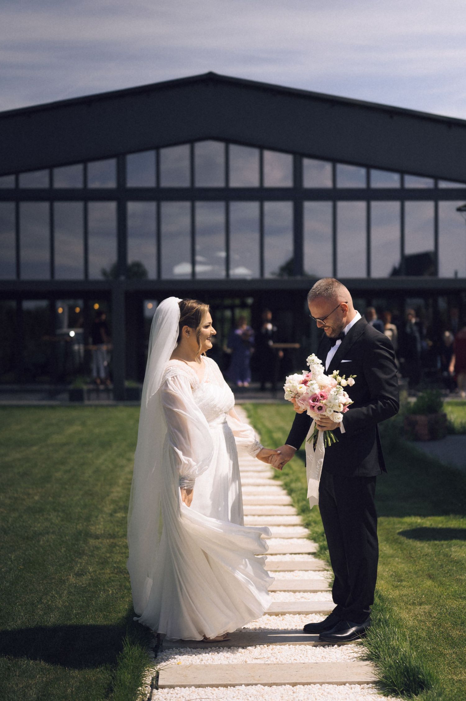 Bride in white gown and groom in black suit exchange vows outdoors, holding a pink bouquet before a modern glass venue.