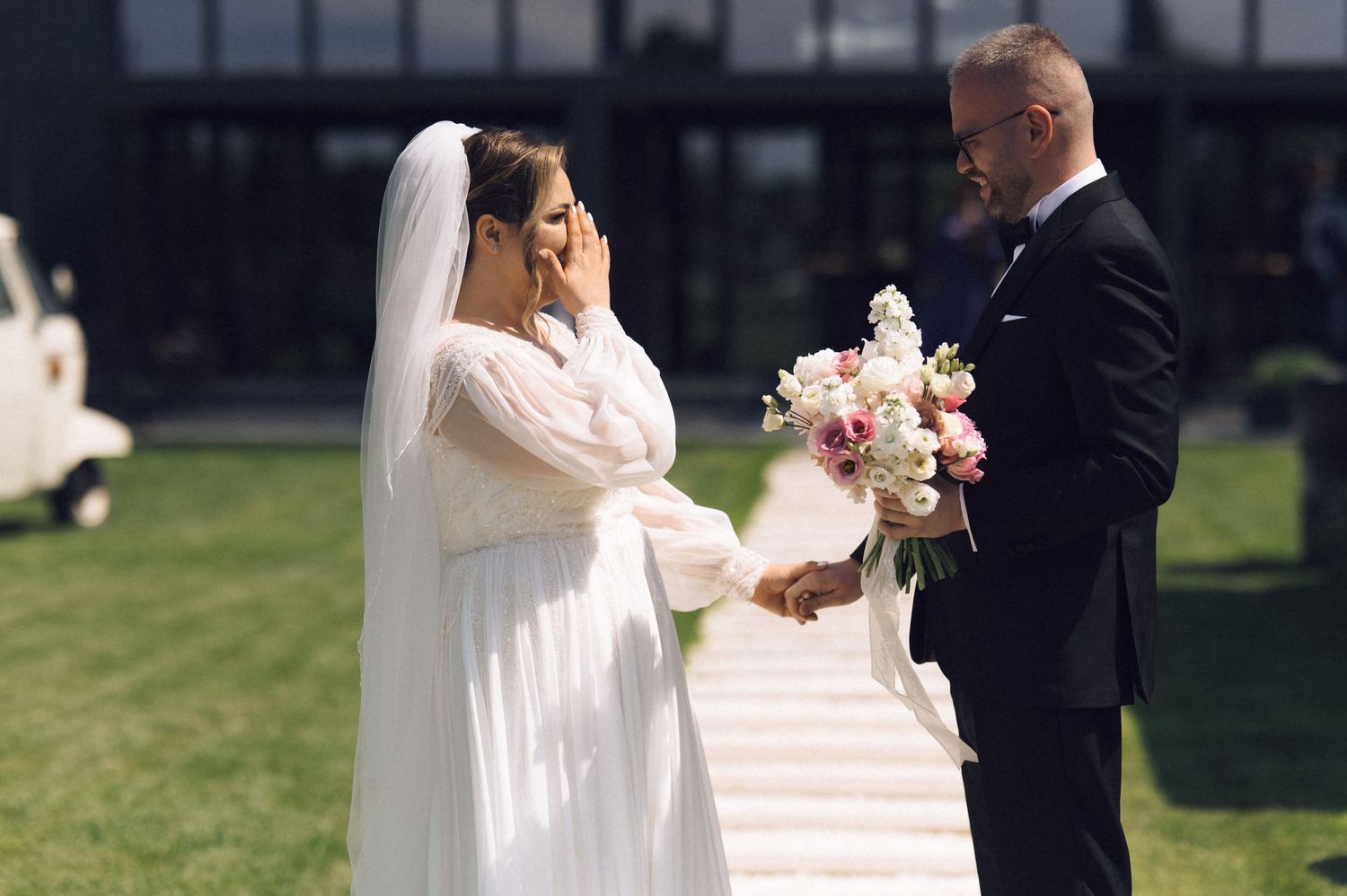 Emotional bride wiping tears while groom holds bouquet of pink and white flowers during outdoor wedding ceremony.