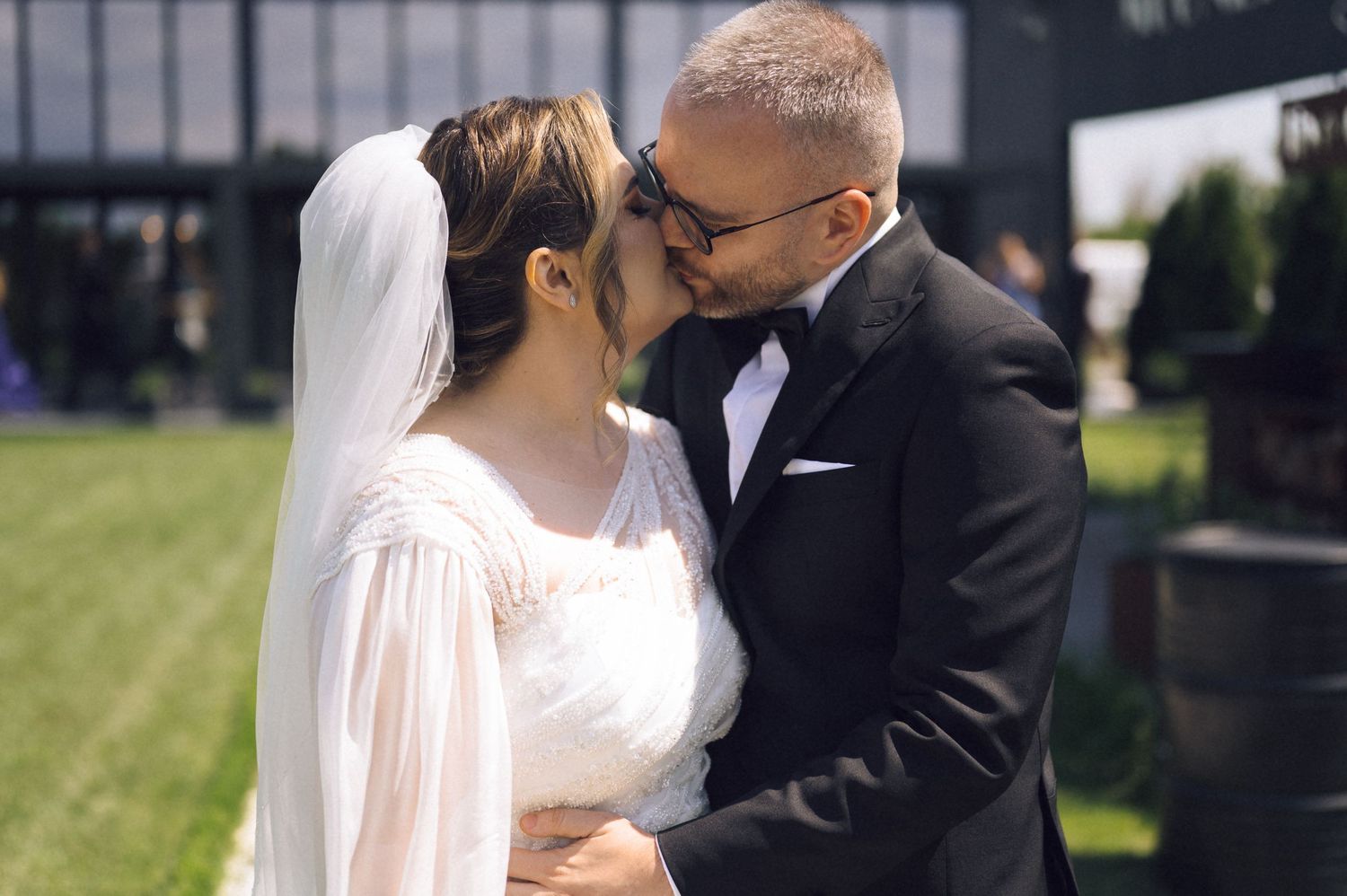 A bride and groom share a romantic first kiss outdoors on their wedding day, surrounded by lush greenery.