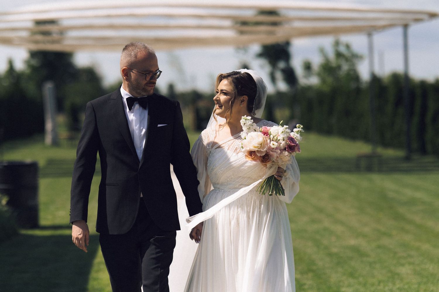 Bride and groom walking outdoors on wedding day, bride holding bouquet in white gown, groom in black tuxedo.